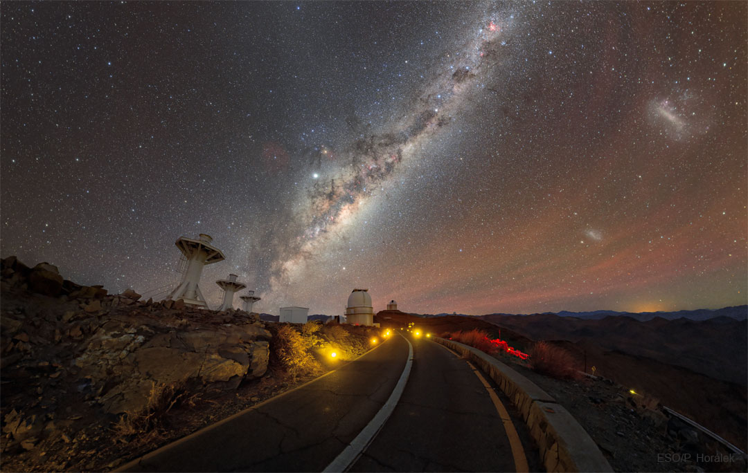 Zdjęcie drogi do Obserwatorium La Silla w Chile. Widoczne są teleskopy oraz gwiazdy, galaktyki, planety oraz poświata niebieska.
Więcej szczegółowych informacji w opisie poniżej.