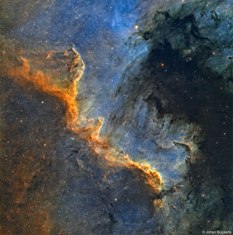 Prezentowane zdjęcie przedstawia Ścianę Łabędzia, postrzępioną wstęgę jasnego gazu oraz ciemnego pyłu na niebieskim tle. Więcej szczegółowych informacji w opisie poniżej.