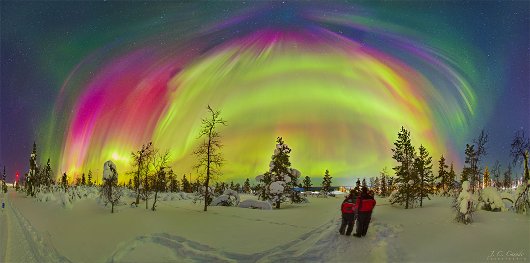 Na zaśnieżonym krajobrazie z gołymi drzewami stoi dwoje ludzi ubranych 
w czerwone płaszcze. Nad nimi widocznych jest wiele barwnych zórz polarnych
w różnych kolorach. W tle zaś widoczne są niektóre gwiazdy. Zobacz opis.
Po kliknięciu obrazka załaduje się wersja
 o największej dostępnej rozdzielczości.