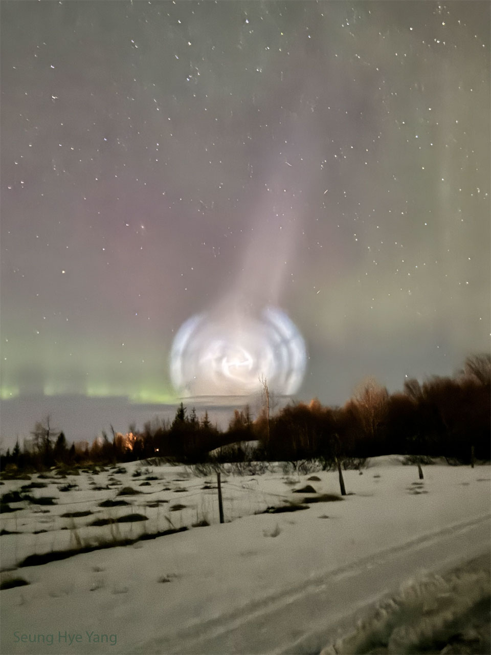 A field of snow is shown, lined with trees along the back.
Above the horizon is an unusual white spiral cloud. 
Stars dot the background, and faint green and red aurora
are also visible. 
Więcej szczegółowych informacji w opisie poniżej.