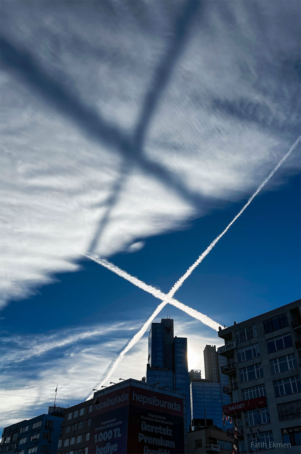 Two airplane contrails, crossing in an X, are shown across
the middle of the image. They are bright white against a dark
blue background. A high cloud deck is seen above the crossing,
sunlit, contrails. A low Sun creates a dark shadow X on the high
while clouds. A row of buildings runs across the lower
part of the image. 
Więcej szczegółowych informacji w opisie poniżej.