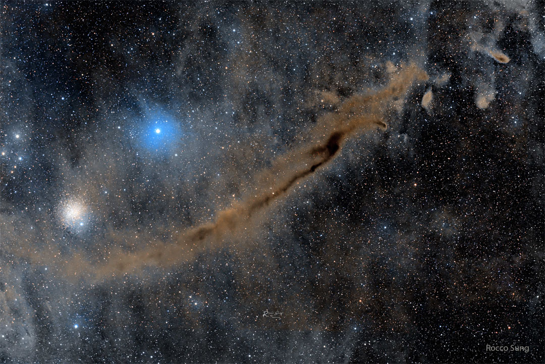 A busy starfield is shown which an elongated brown 
nebula running diagonally from the lower left to the 
upper right. A bright blue star and a star cluster 
appear above the nebula. 
Więcej szczegółowych informacji w opisie poniżej.