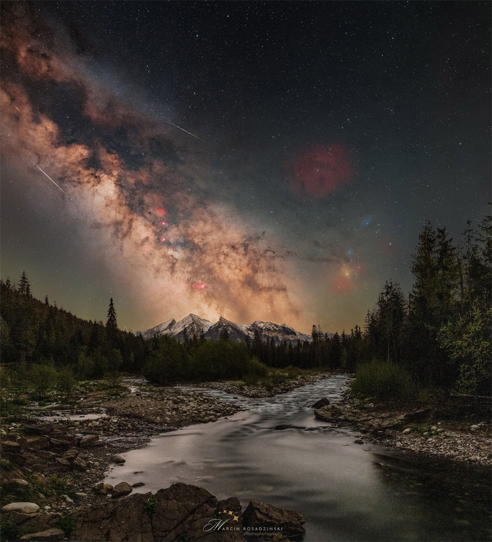 A starry sky is shown with the busy central band of 
our Milky Way Galaxy showing diagonally from the upper left.
Mountains are on the horizon, with trees and a stream running
up from the foreground. 
Więcej szczegółowych informacji w opisie poniżej.