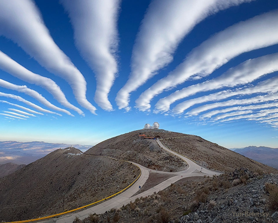 A series of white parallel clouds are seen going off
into the distance in a background blue sky. In the foreground
is a hill with two domes at the top. 
Więcej szczegółowych informacji w opisie poniżej.