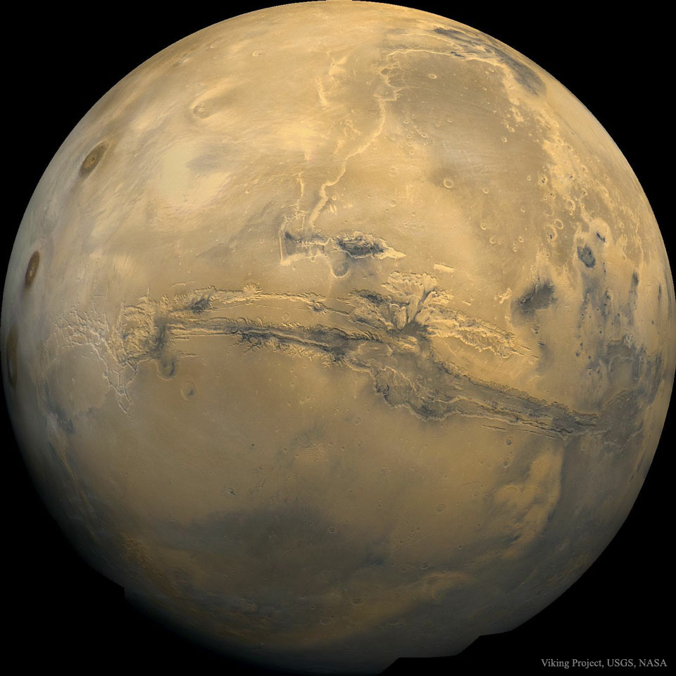 A picture of Mars is shown as a large orange globe.
Across the center of the planet a long canyon is visible. 
Więcej szczegółowych informacji w opisie poniżej.