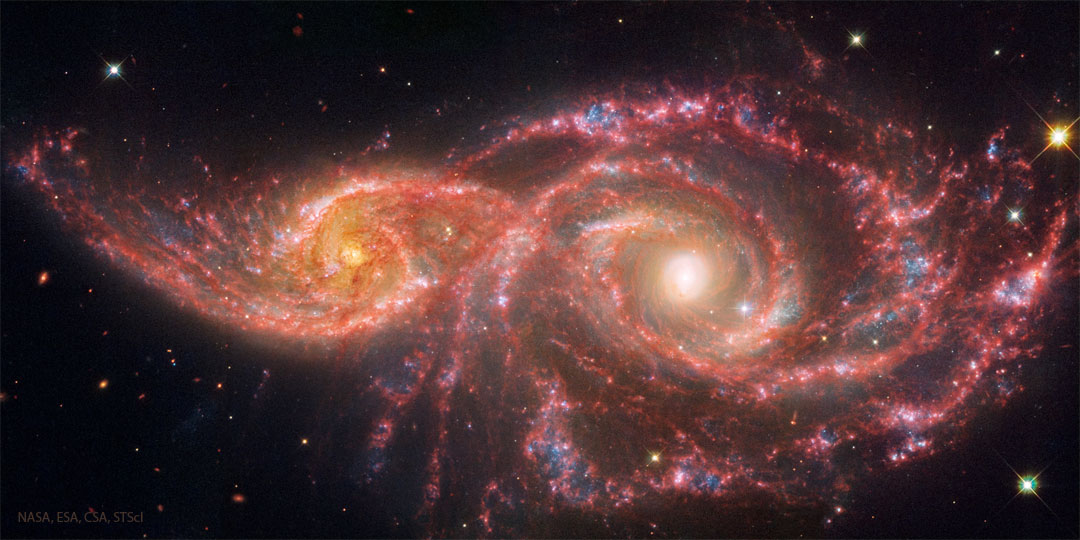 Two spiral galaxies are pictured on the left and right. 
They galaxy on the left is smaller. Both show red lanes of
dust in their spiral arms. 
Więcej szczegółowych informacji w opisie poniżej.