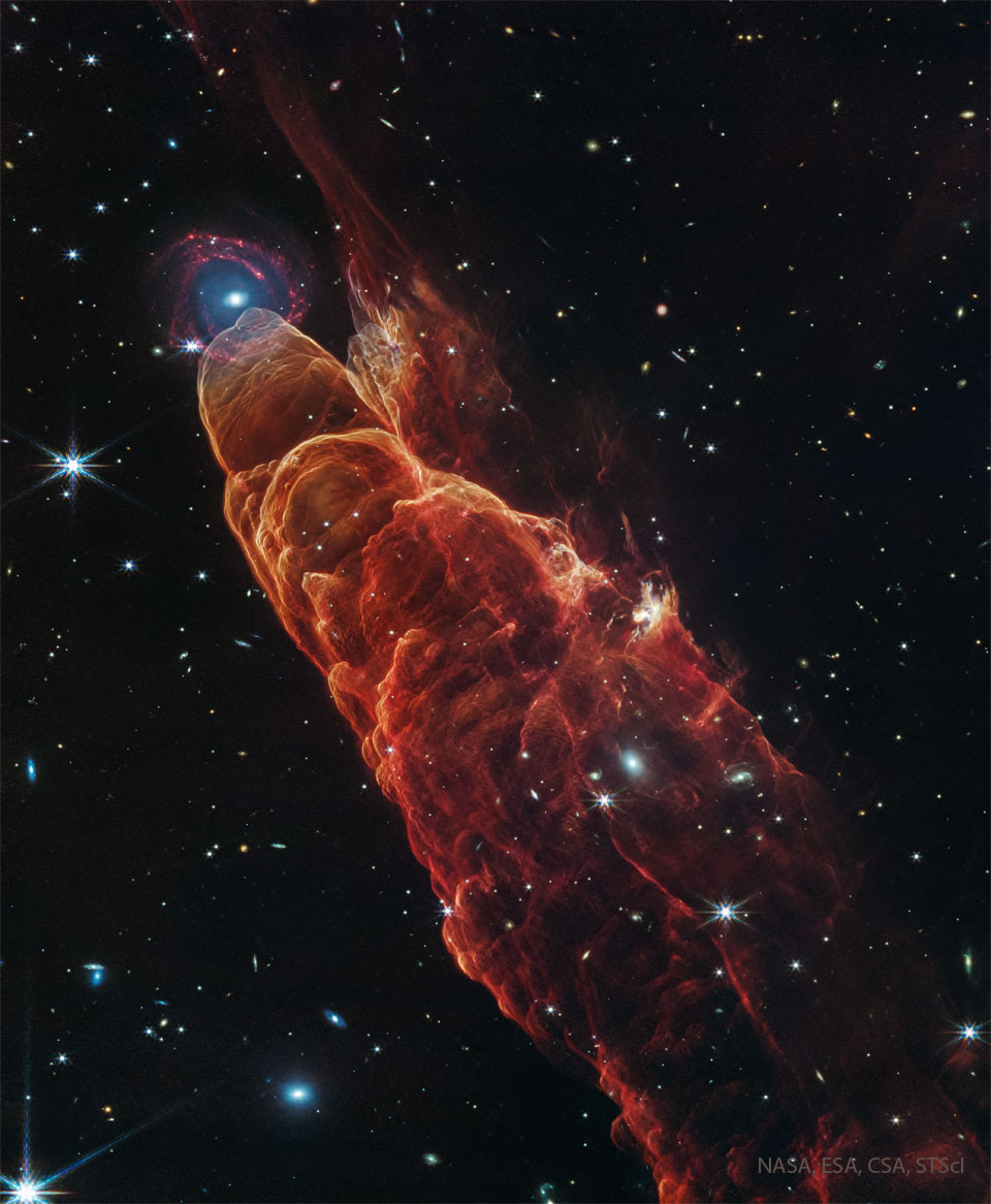 A dark starfield is dominated by a red cone-like
nebula with its base on the bottom right and extending
diagonally through the image. At the peak of the cone
toward the upper left is a background spiral galaxy.
Więcej szczegółowych informacji w opisie poniżej.