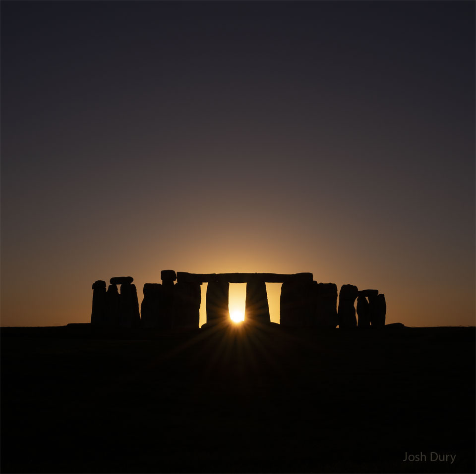 Na zdjęciu na czystym niebie ponad polem zachodzi
Słońce. Na polu znajdują się słynne kamienie Stonehenge.
Samo Słońce prześwituje w centrum przez układ kamieni.
Zobacz opis. Po kliknięciu obrazka załaduje się wersja
 o&nbsp;największej dostępnej rozdzielczości.