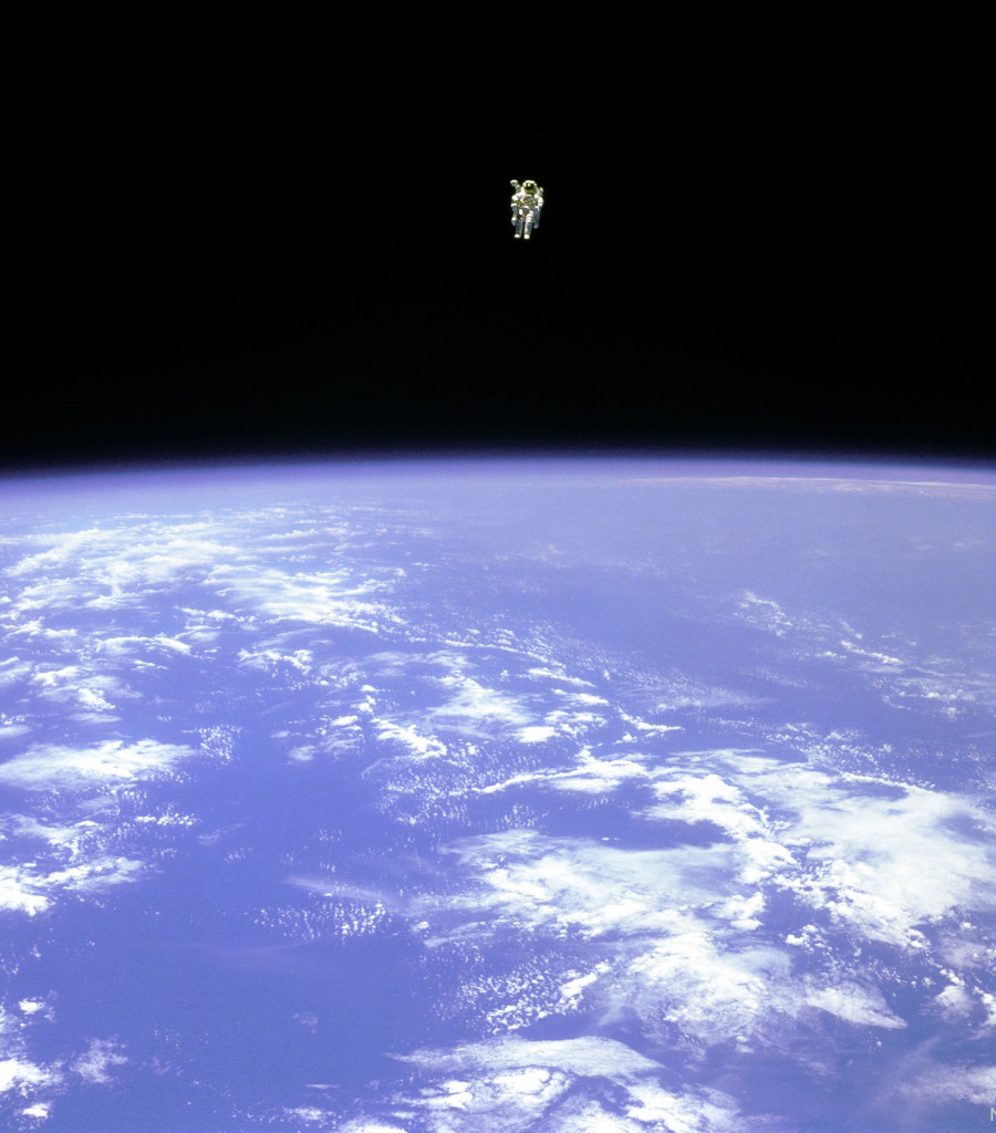 Na tle ciemnej przestrzeni kosmicznej unosi się astronauta. 
W&nbsp;dolnej części zdjęcia widać jasną, niebieską Ziemię oraz jej białe chmury.
Więcej szczegółowych informacji w opisie poniżej.