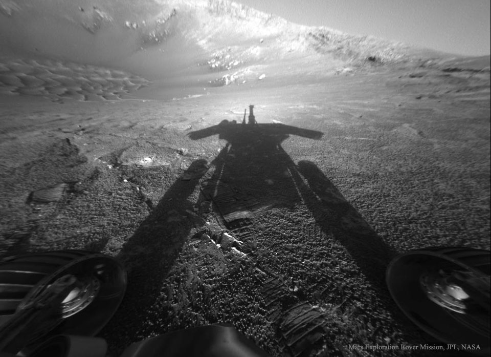 A black and white image shows an unusual landscape with a
bright ridge across the top and texture like small sand dunes. 
The shadow of something unusual dominates the image center. 
Wheels are visible at the bottom of the image. near the middle. 
Więcej szczegółowych informacji w opisie poniżej.