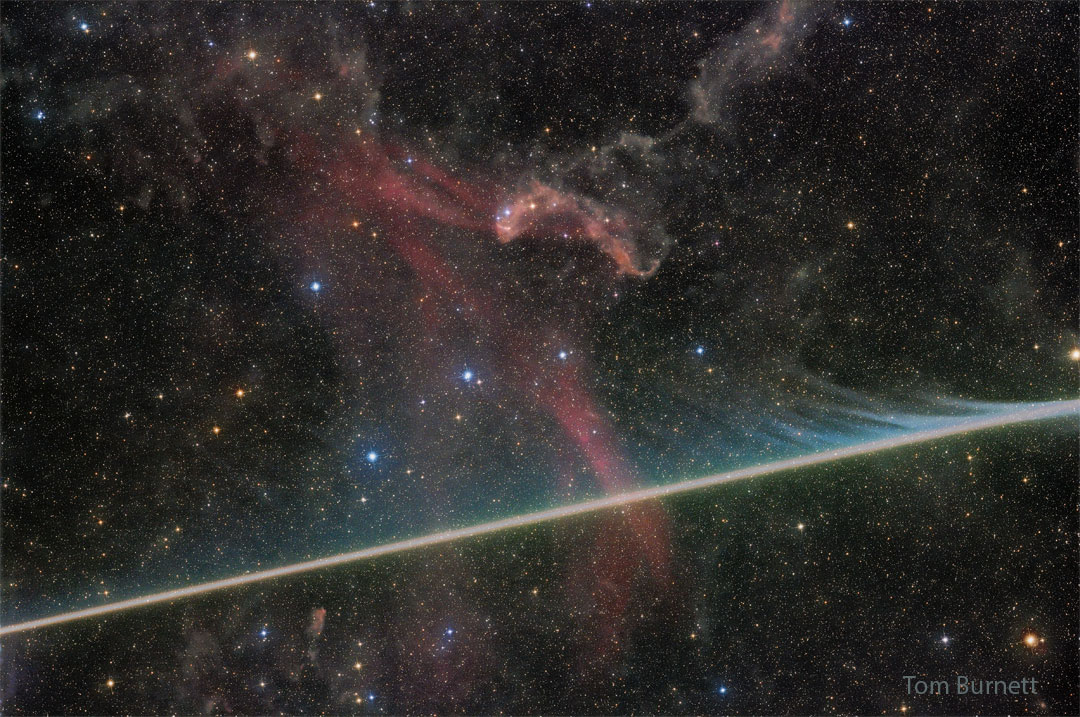 A dark starfield has a faint red nebula near 
the center. A bright streak crosses the image 
horizontally from the lower left, filled with faint
evaporating wisps in many colors. 
Więcej szczegółowych informacji w opisie poniżej.